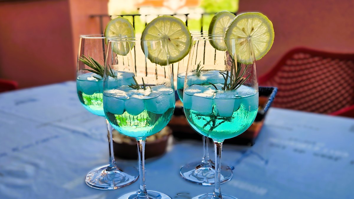 Das BIld zeigt vier Gläser Venturo Spritz mit der charakteristischen blaugrünen Farbe, auf den Gläsern stecken Zitronenscheiben, im Drink schwimmen Eiswürfel und Rosmarinzweige
