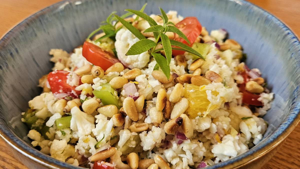 Karfiol-Rohkostsalat in baluer Bowl: Die Zutaten sind gut zu erkennen - Blumenkohl gerieben, Tomaten- und Paprikastücke, Orangenzeste und -stücke, Pinienkerne und Verbenenspitzen