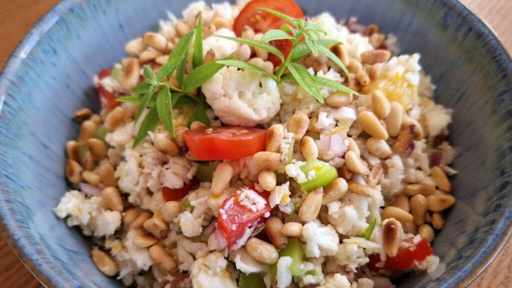 Karfiol-Rohkostsalat in Nahaufnahme: Die Zutaten sind gut zu erkennen - Blumenkohl gerieben, Tomaten- und Paprikastücke, Orangenzeste und -stücke, Pinienkerne und Verbenenspitzen, angerichtet in einer hellblauen Bowl